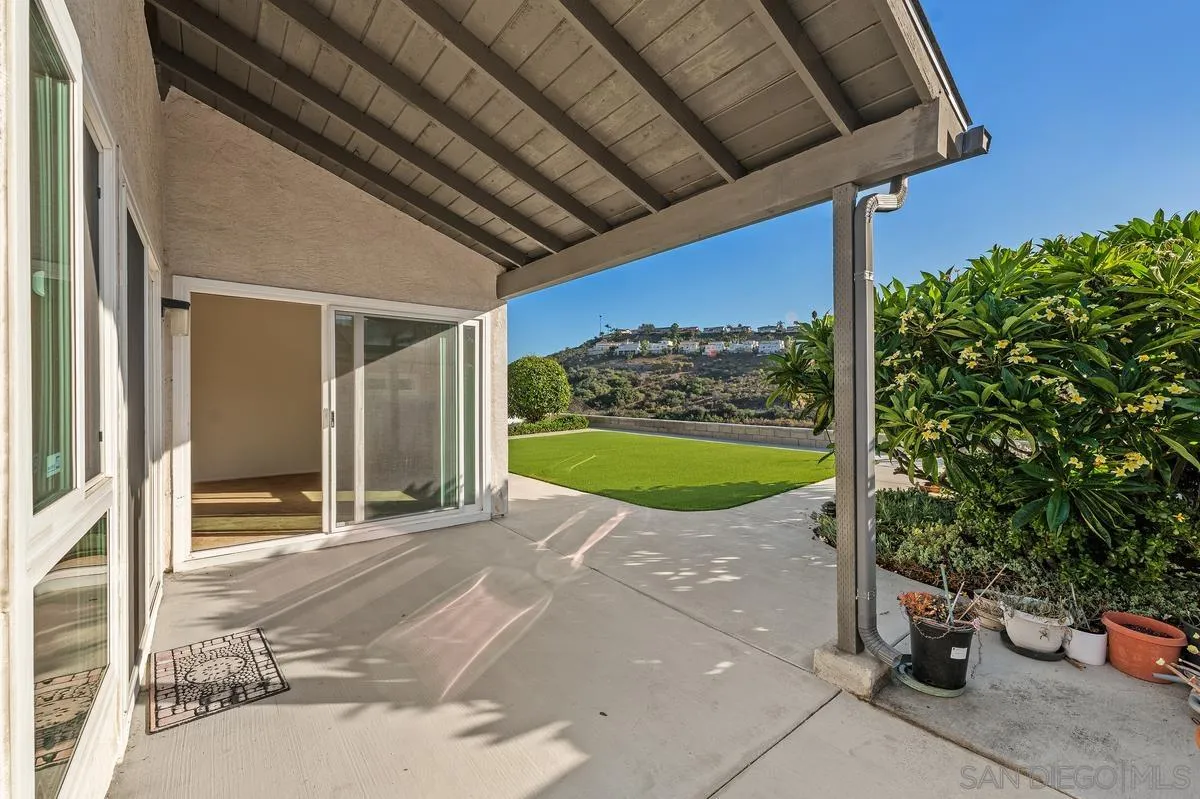 Covered patio with sliding glass doors, potted plants, lawn, and distant hillside homes.