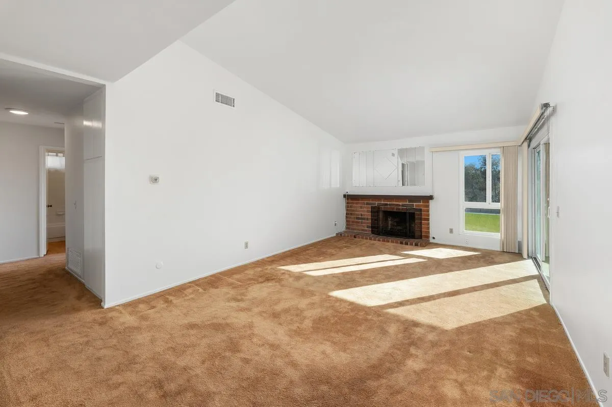 Empty living room with brick fireplace, tan carpet, and sunlit sliding glass doors.