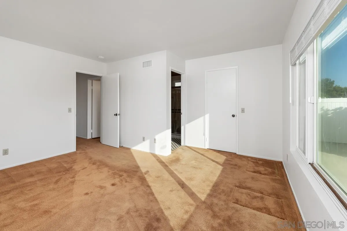 Empty bedroom with tan carpet and sunlight streaming through a large window.