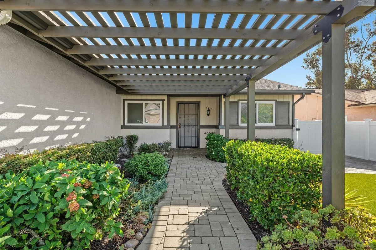 Home entrance beneath a wooden pergola, with a paved path and landscaped shrubs.