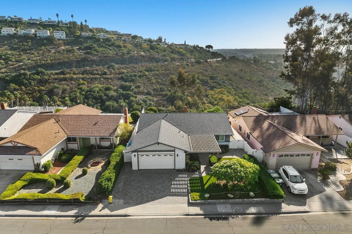 Aerial view of a single-story home with a paved driveway and hillside neighborhood behind it.