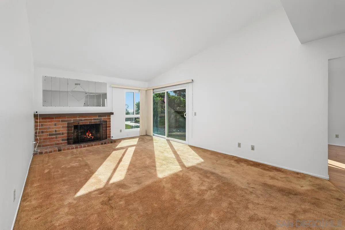 Empty living room with tan carpet, brick fireplace, and sliding glass doors opening to greenery.