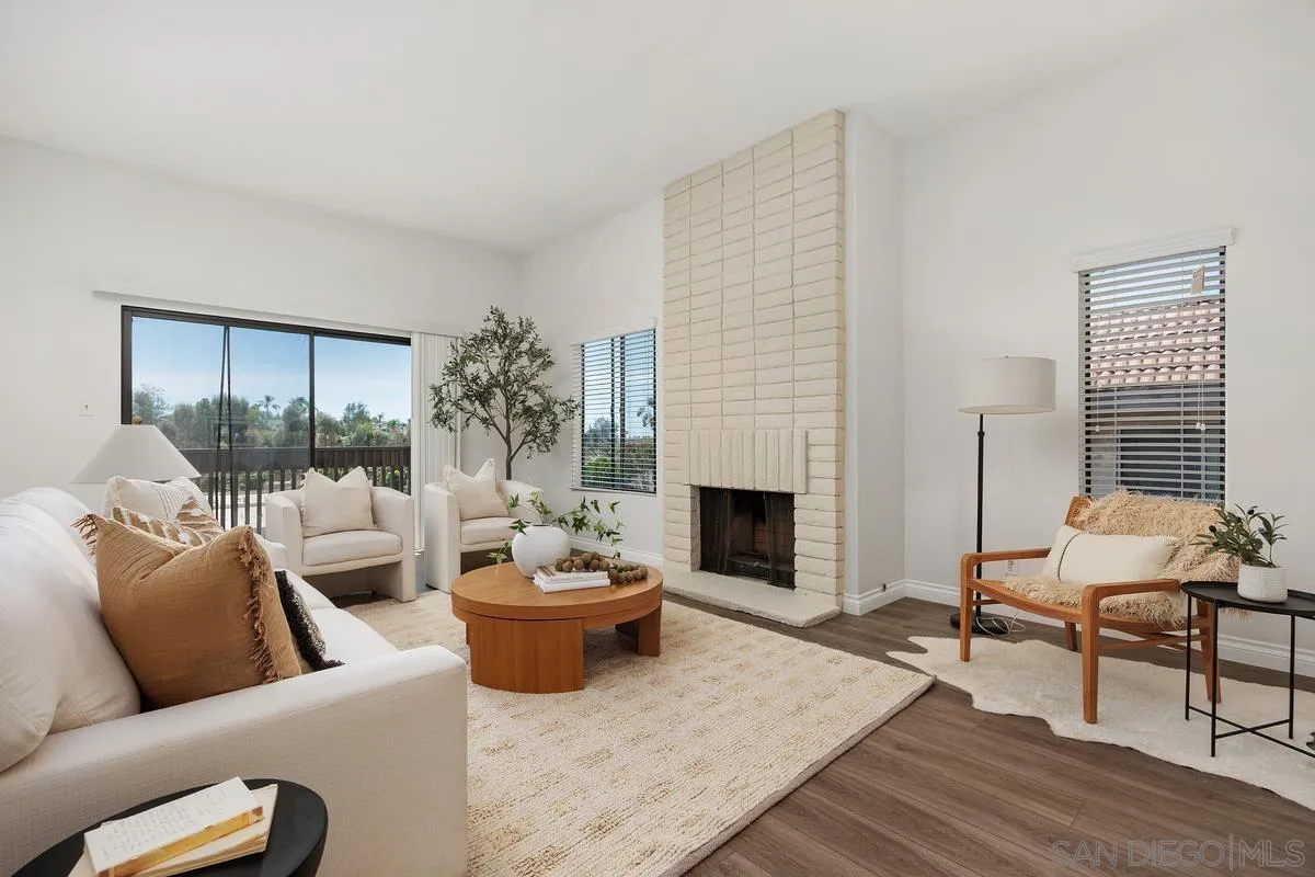 Staged living room with a tall white brick fireplace, white seating, and large windows overlooking greenery.