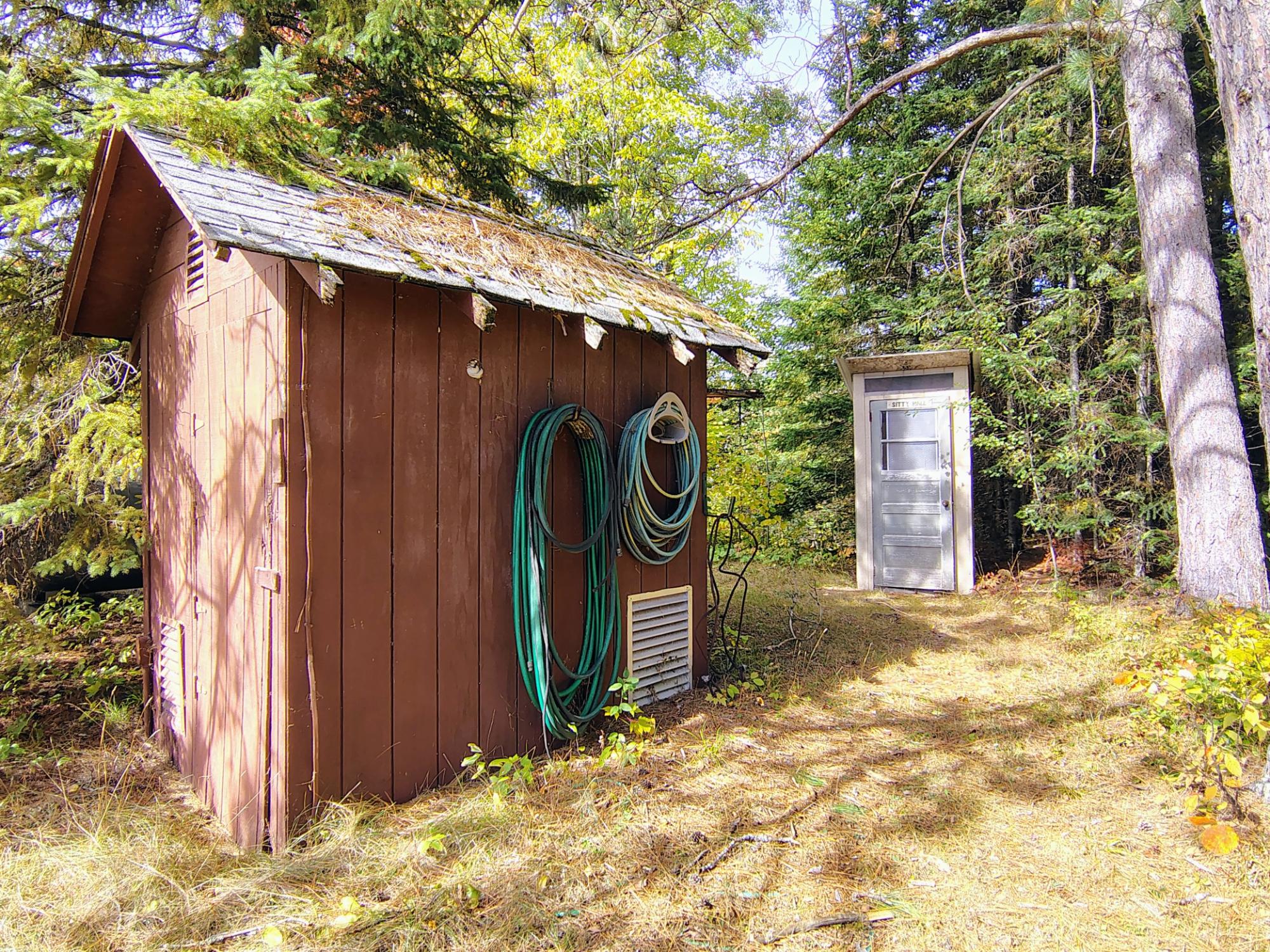 Wooden utility shed with coiled hoses beside a small detached toilet building in the woods.