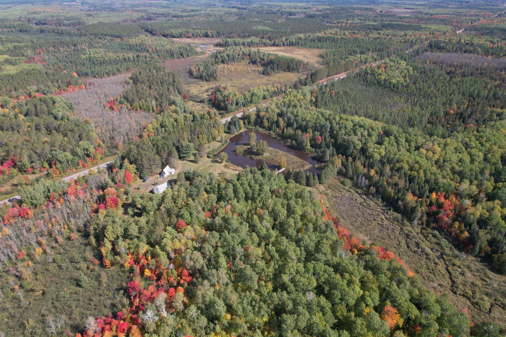 Aerial view of a wooded property with a pond, small bridge, and buildings amid autumn foliage.
