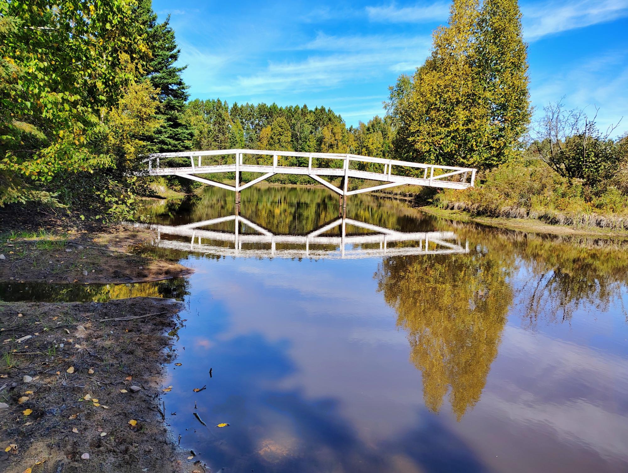 White footbridge reflected in calm water, with autumn trees lining the banks.