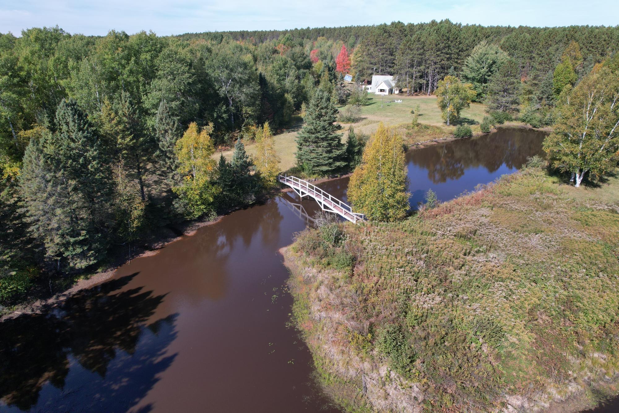 Wooded property with a pond, white footbridge, and house in the distance.
