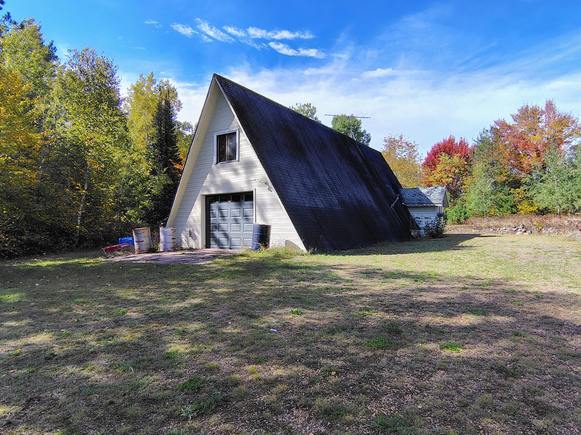 White A-frame building with a garage door in a grassy clearing surrounded by autumn trees.