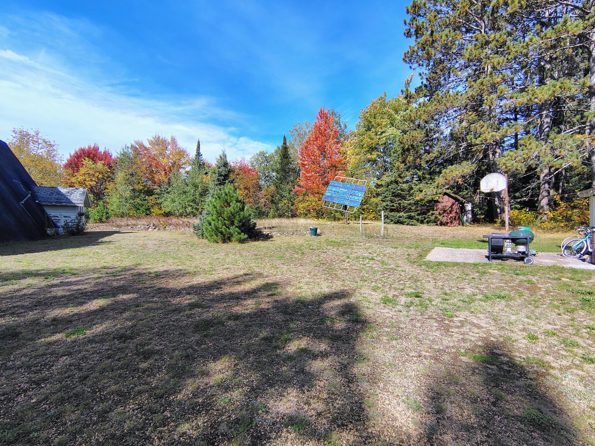 Grassy backyard bordered by autumn trees, with solar panels, a small outbuilding, and a basketball hoop.