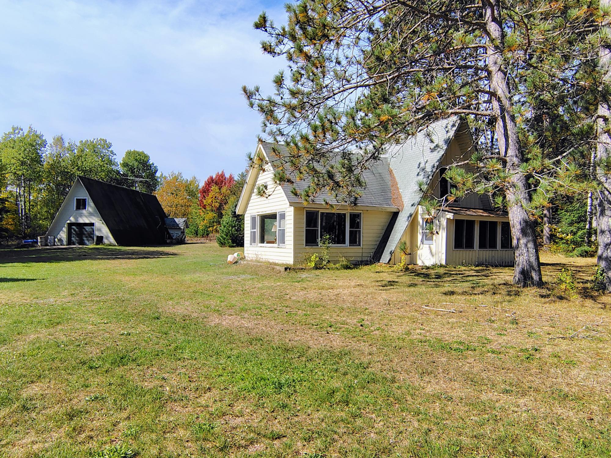 Light-colored A-frame house and detached outbuilding on a grassy lawn beneath mature pine trees.