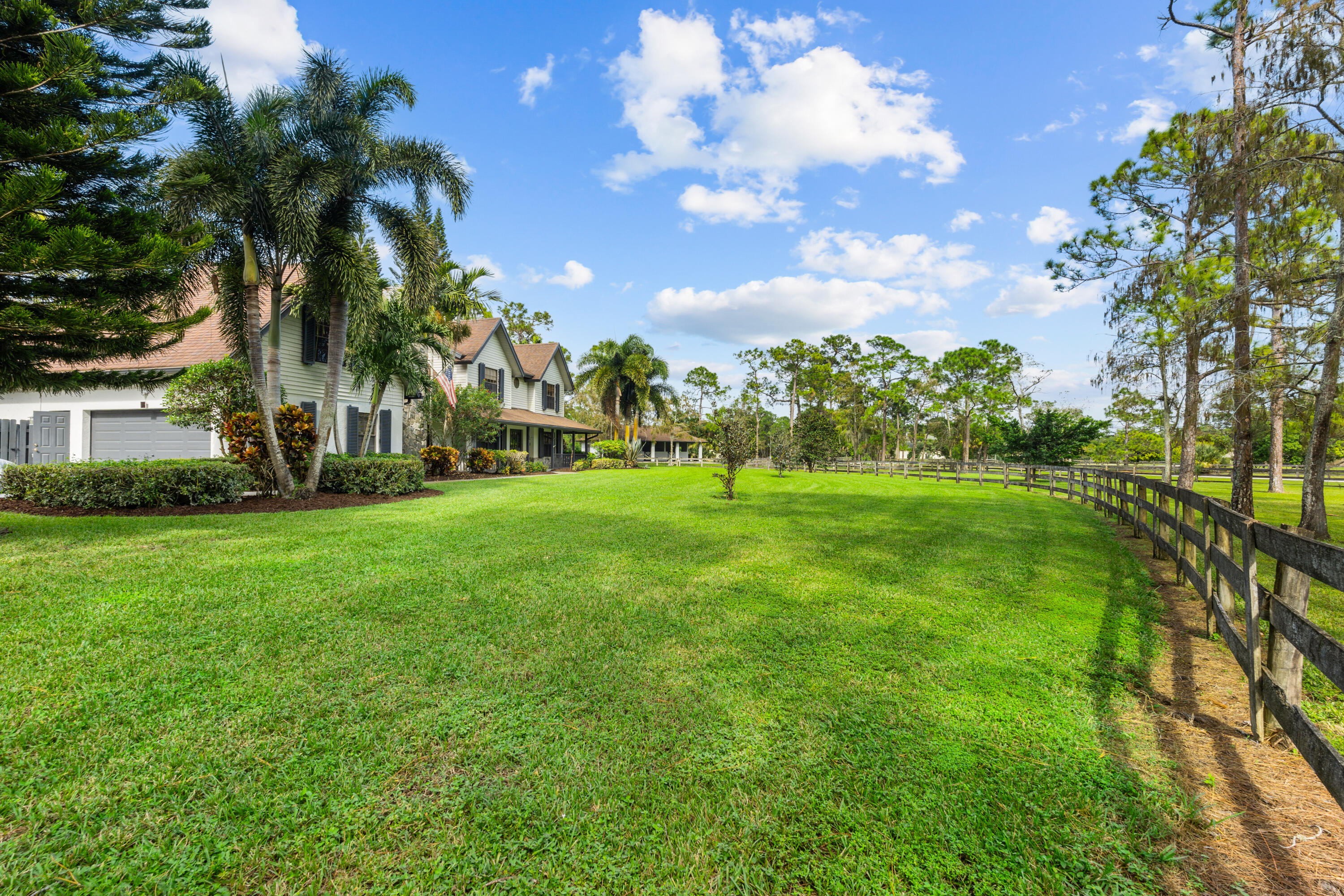 Large two-story home and garage beside an expansive lawn bordered by a dark wooden fence and palm trees.