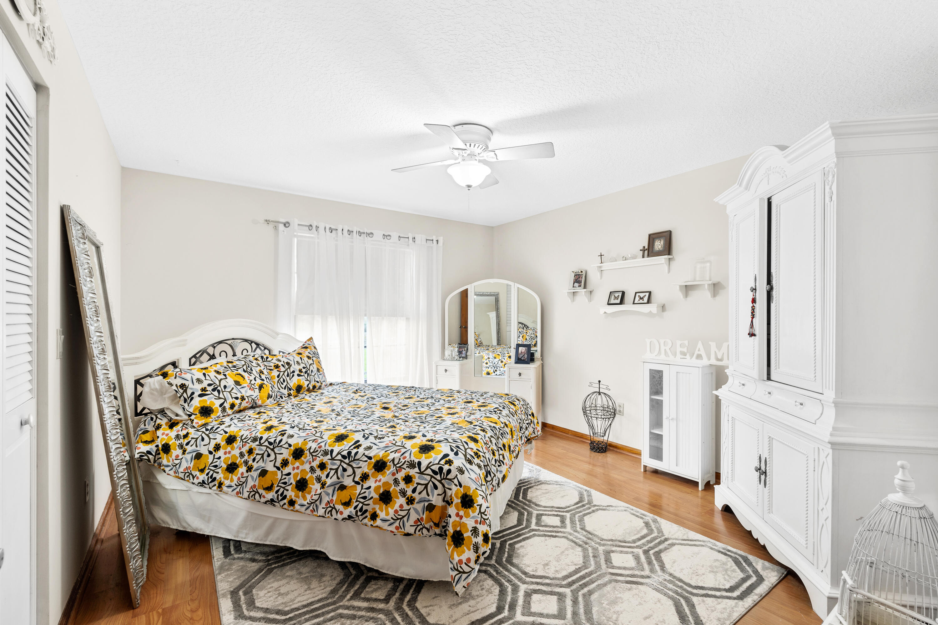 Furnished bedroom with floral bedding, a large geometric rug, mirrored vanity, and ornate white wardrobes.