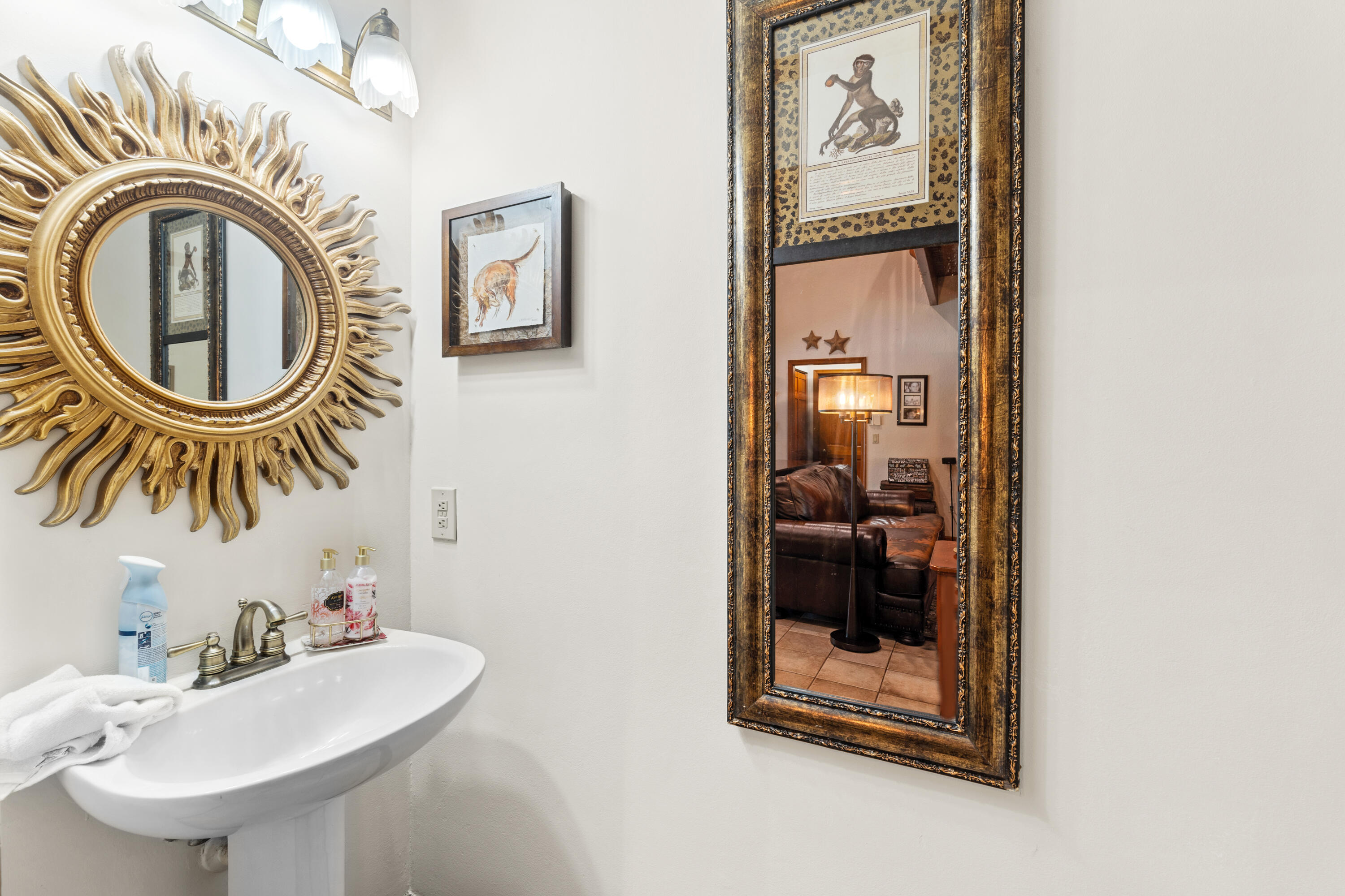Bathroom pedestal sink with brass faucet, sunburst mirror, and ornate mirror reflecting a sitting area.
