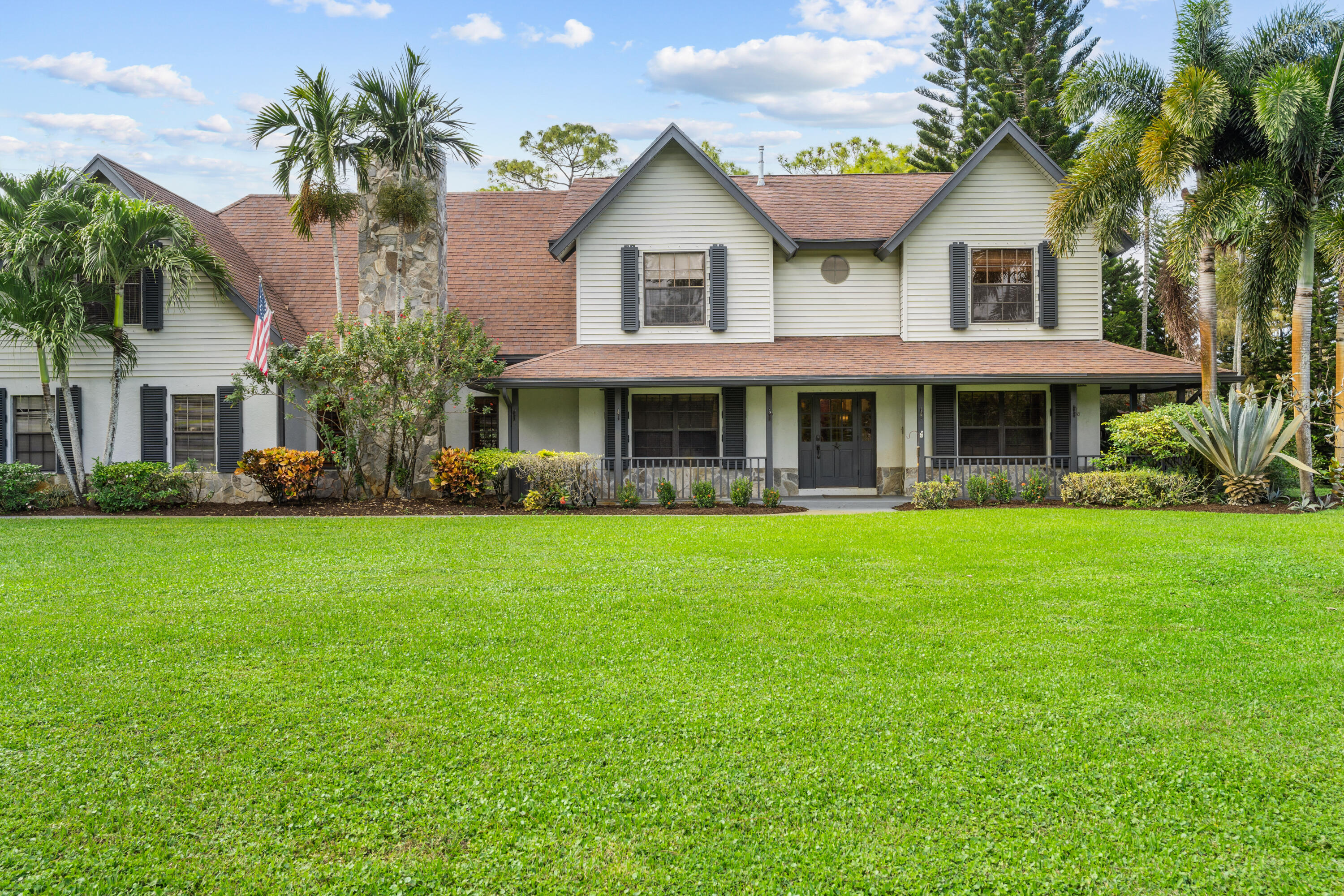 Two-story white house with a covered porch, stone chimney, palm trees, and broad green lawn.