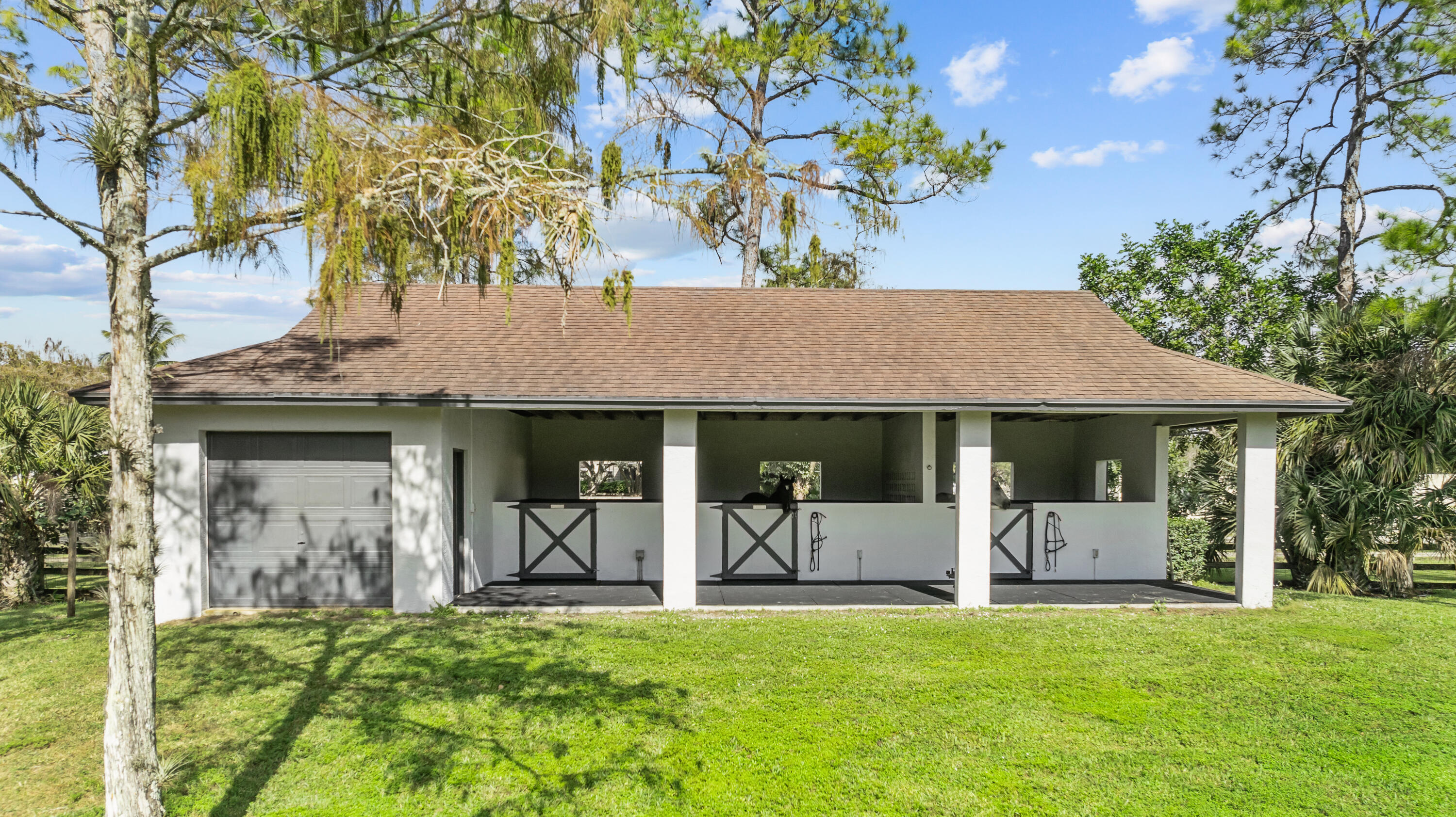 White equestrian stable with open stalls, a gray garage door, and a broad shingle roof.