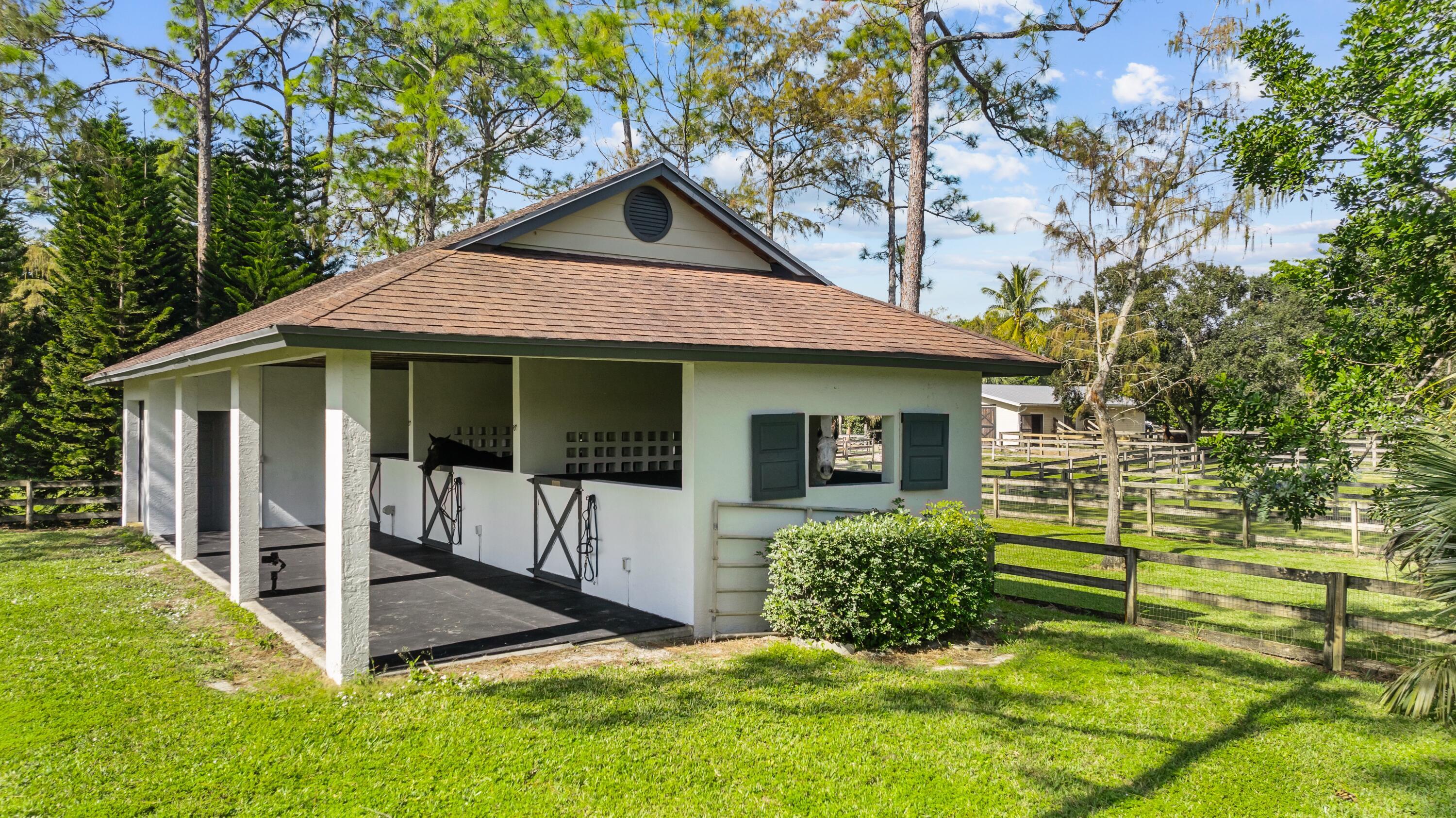 White equestrian stable with open stalls, horses, and fenced paddocks.