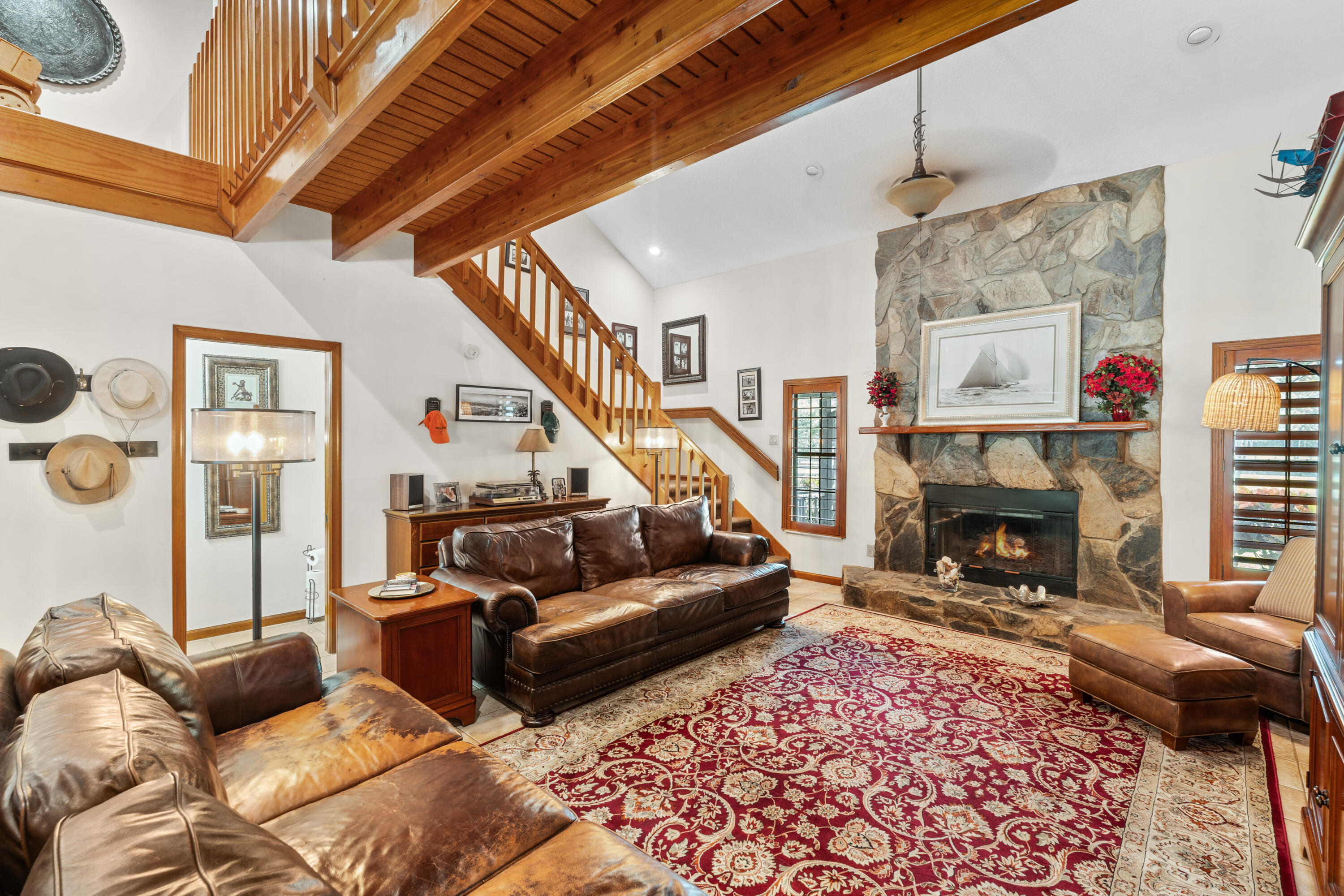 Living room with leather sofas, a stone fireplace, and a dramatic exposed-wood balcony and staircase.