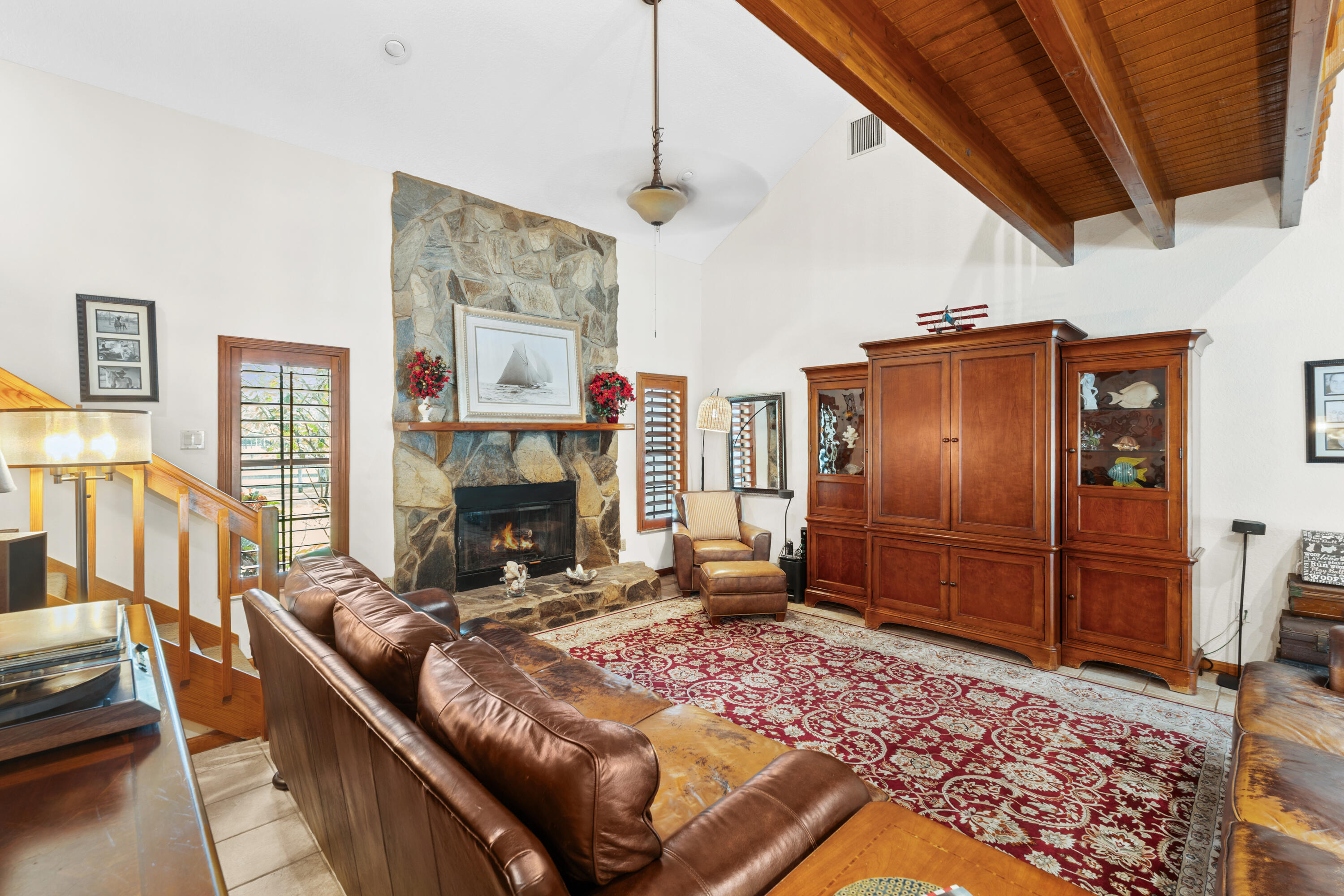 Living room with a stone fireplace, exposed wood ceiling beams, leather seating, and a red patterned rug.
