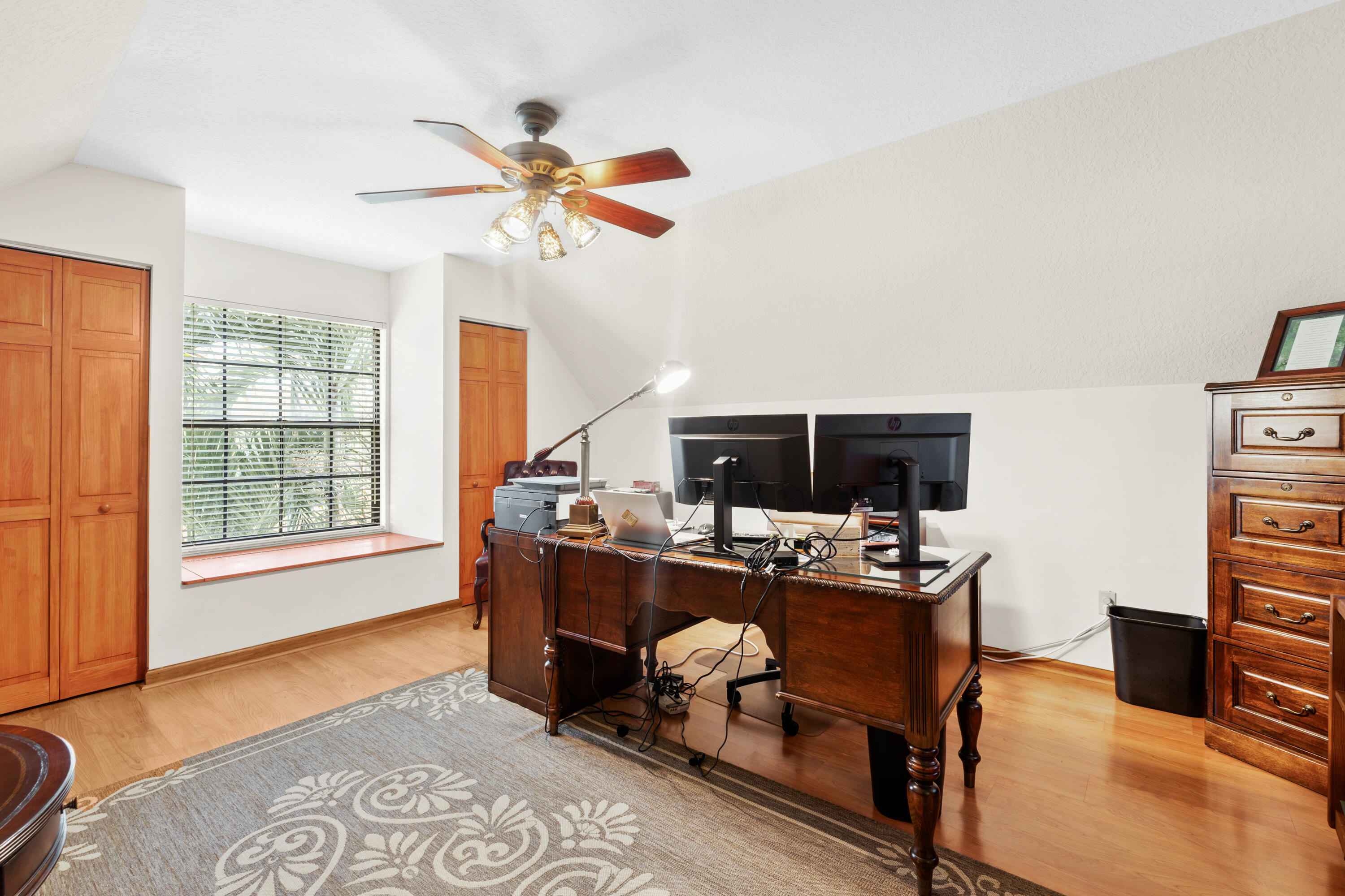 Home office with a dark wood desk, dual monitors, hardwood floors, and a window with blinds.