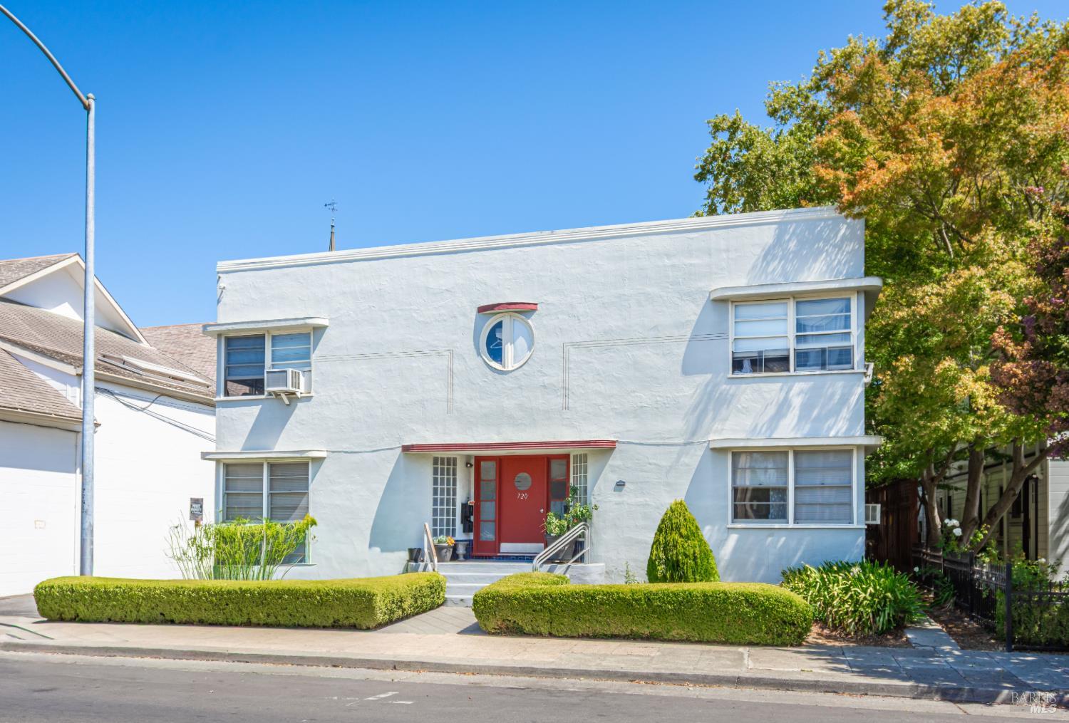 Pale blue building with a round window, red entrance door, and neatly trimmed hedges.