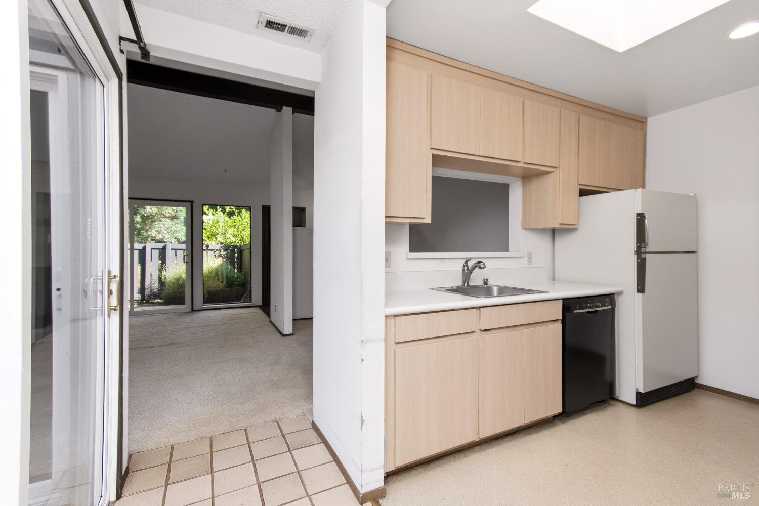 Kitchen with light wood cabinets and white appliances opening to a carpeted room with garden-facing sliding doors.