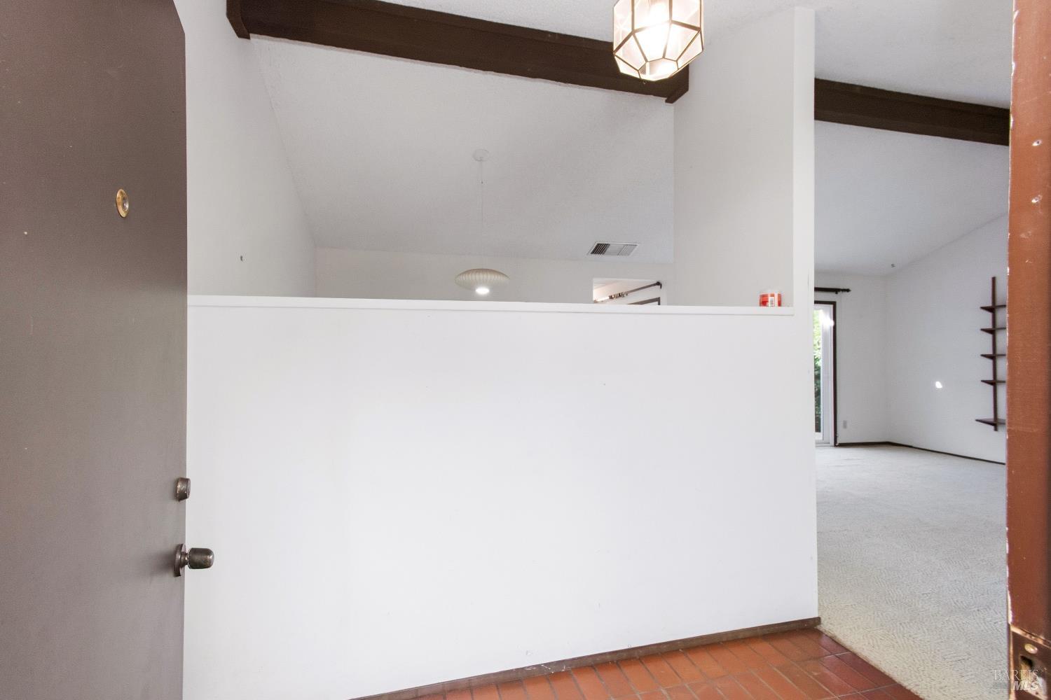 Empty entryway with terracotta tile, a white half-wall, dark ceiling beams, and a carpeted room beyond.