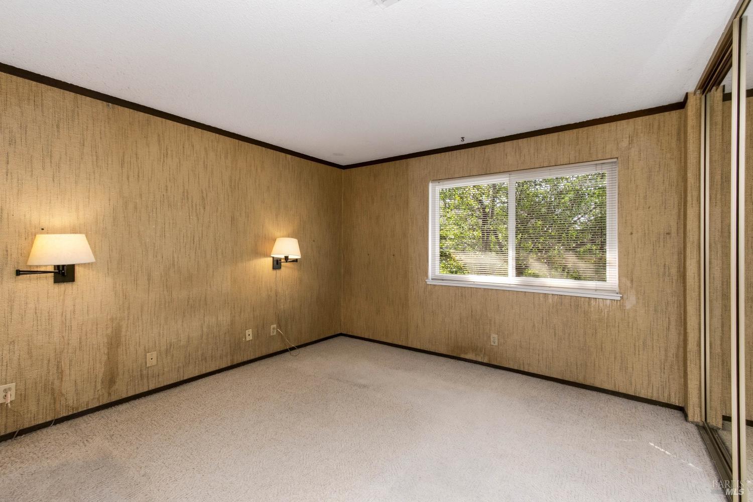 Empty bedroom with tan textured walls, beige carpet, wall sconces, and mirrored closet doors.