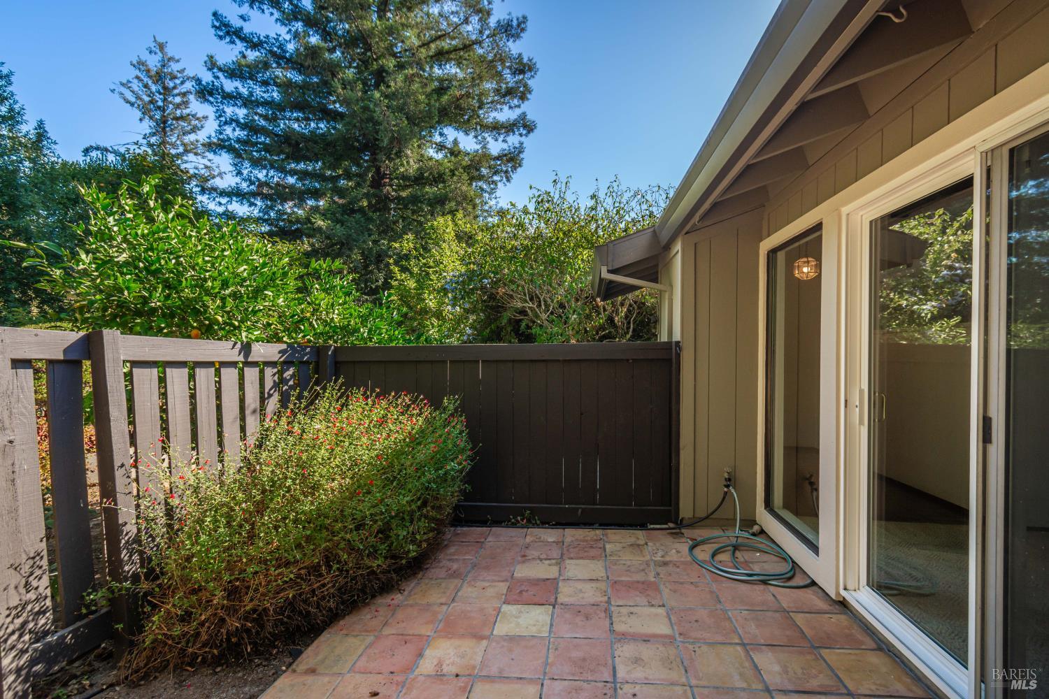 Fenced patio with multicolored tile paving, leafy shrubs, and sliding glass doors.