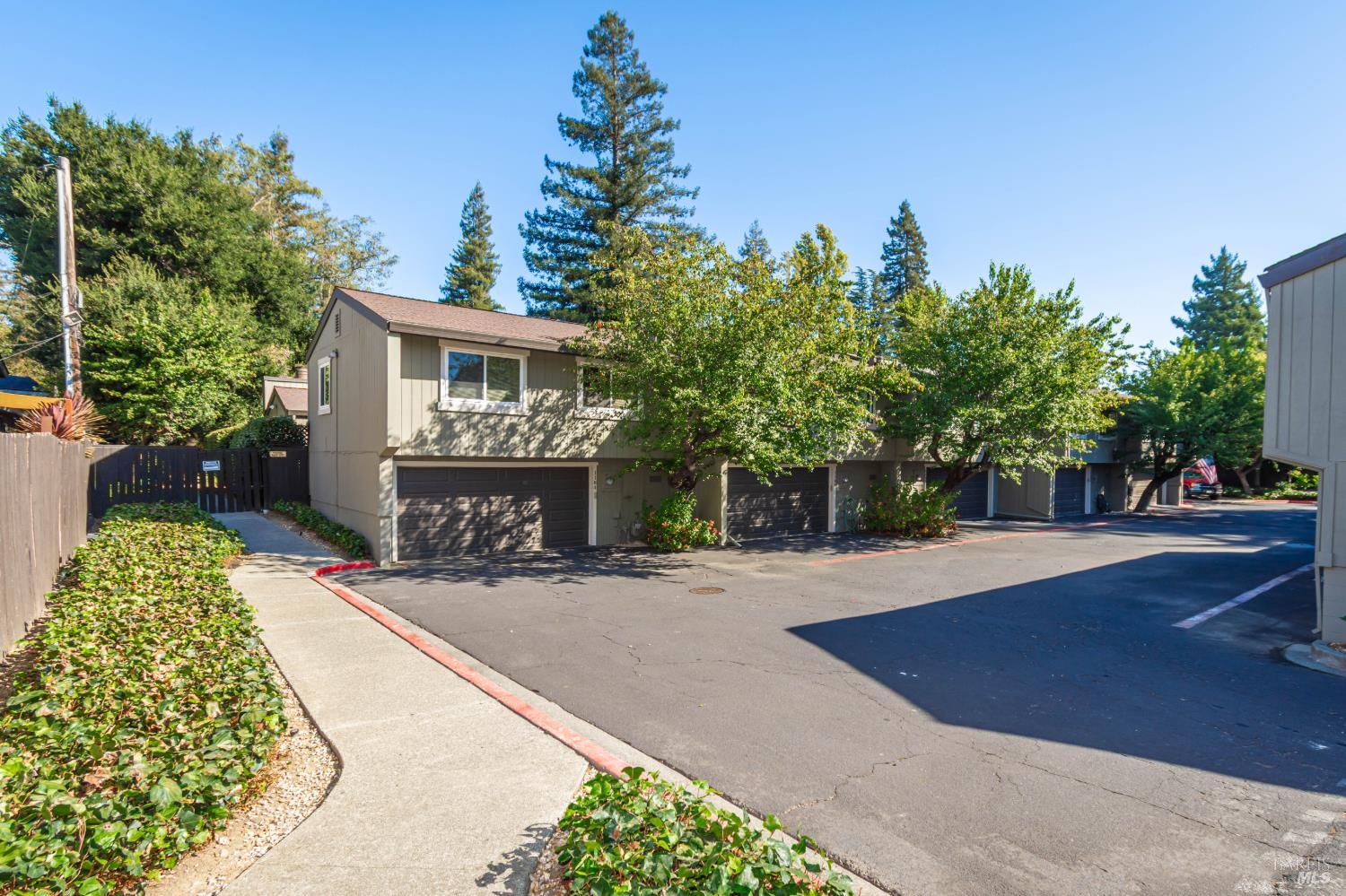 Two-story beige residence with multiple garage doors, leafy landscaping, and a paved parking area.