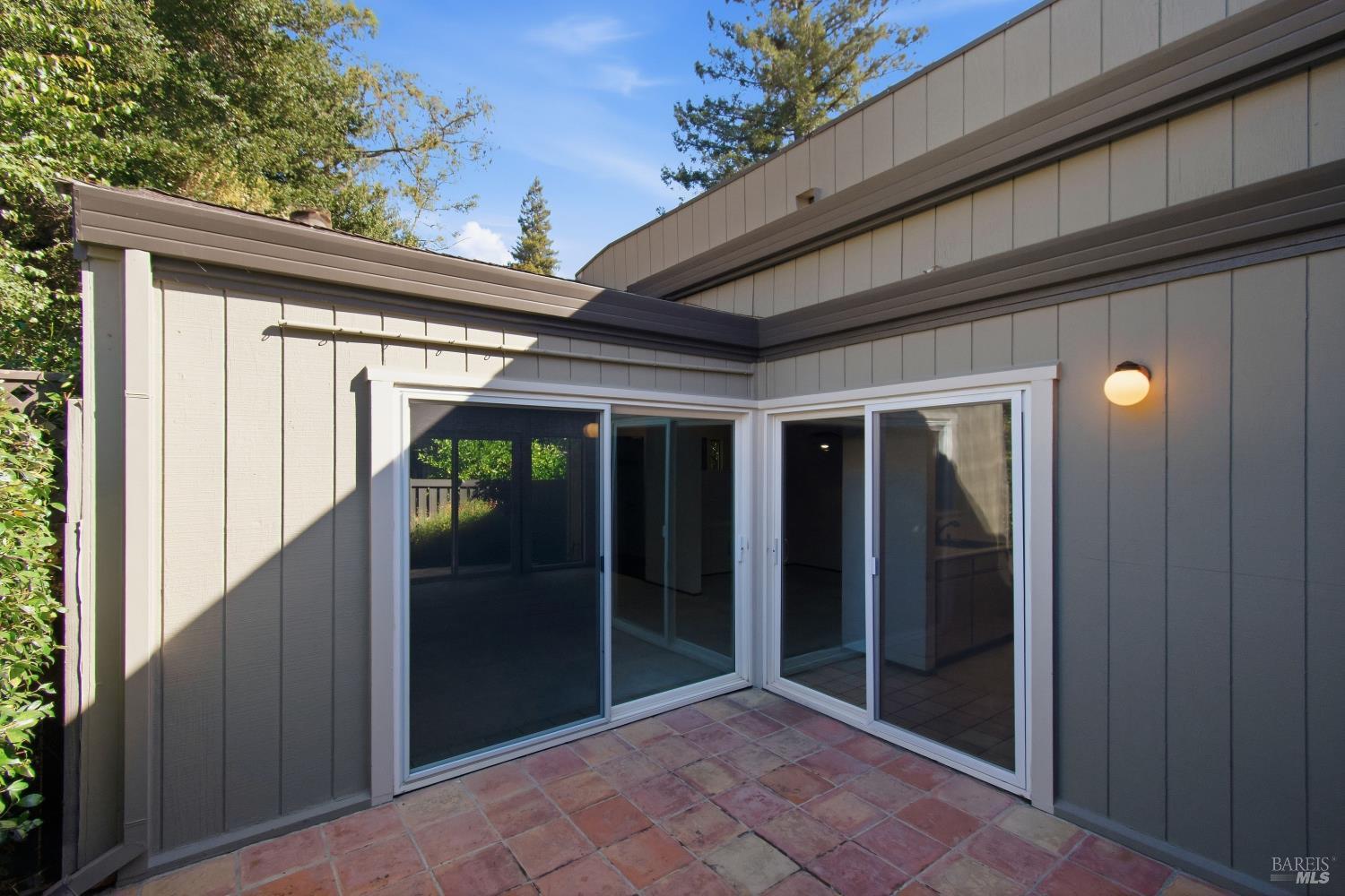 Tiled patio with large sliding glass doors set into gray siding, bordered by trees.