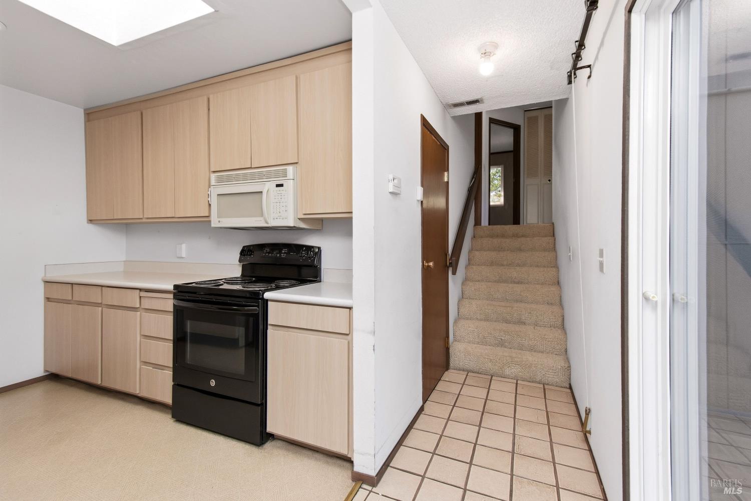 Kitchen with light wood cabinets and black range beside a tiled entry and carpeted staircase.