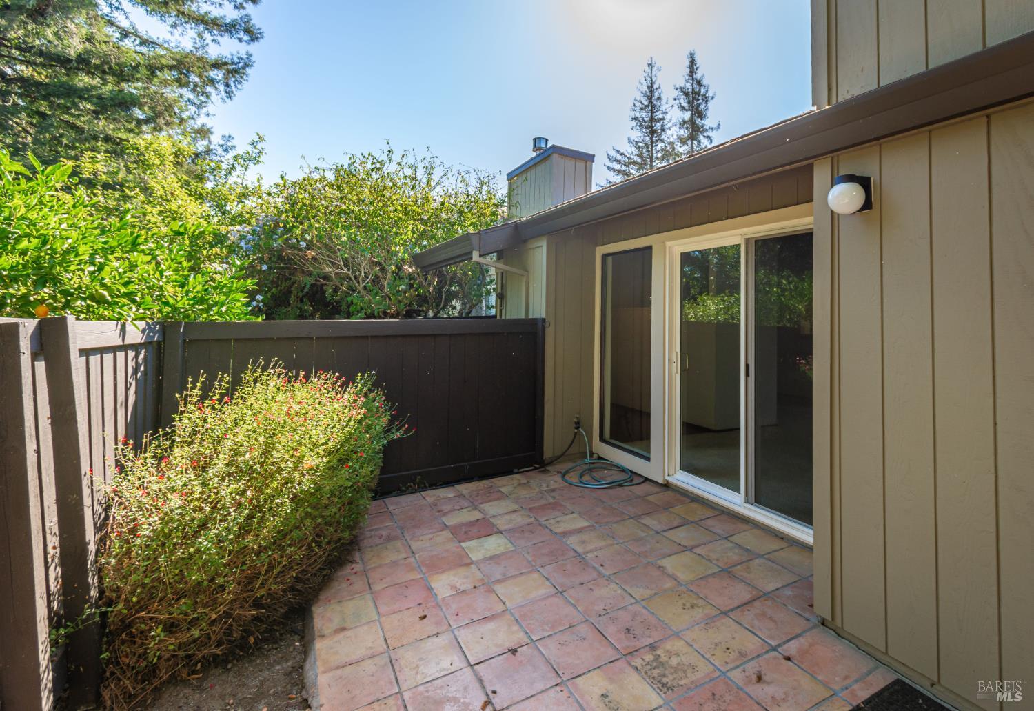 Fenced patio with multicolored tile flooring, shrubs, and a sliding glass door.