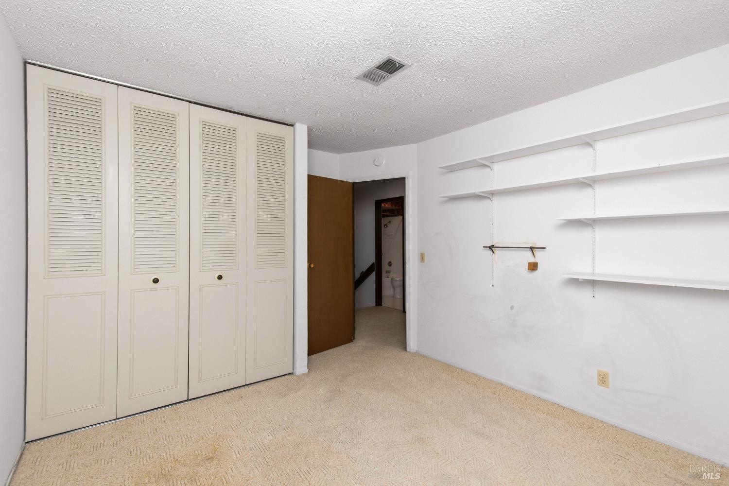 Empty carpeted room with louvered closet doors, wall shelving, and an open bathroom doorway.
