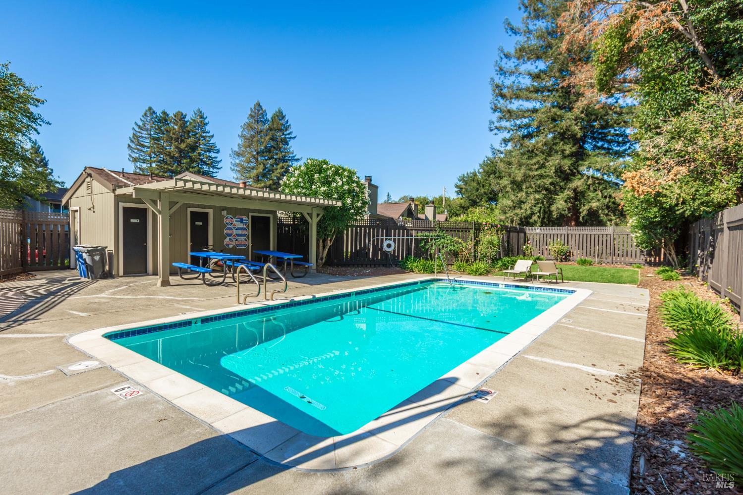 Outdoor swimming pool beside a pergola-covered picnic area and fenced courtyard.