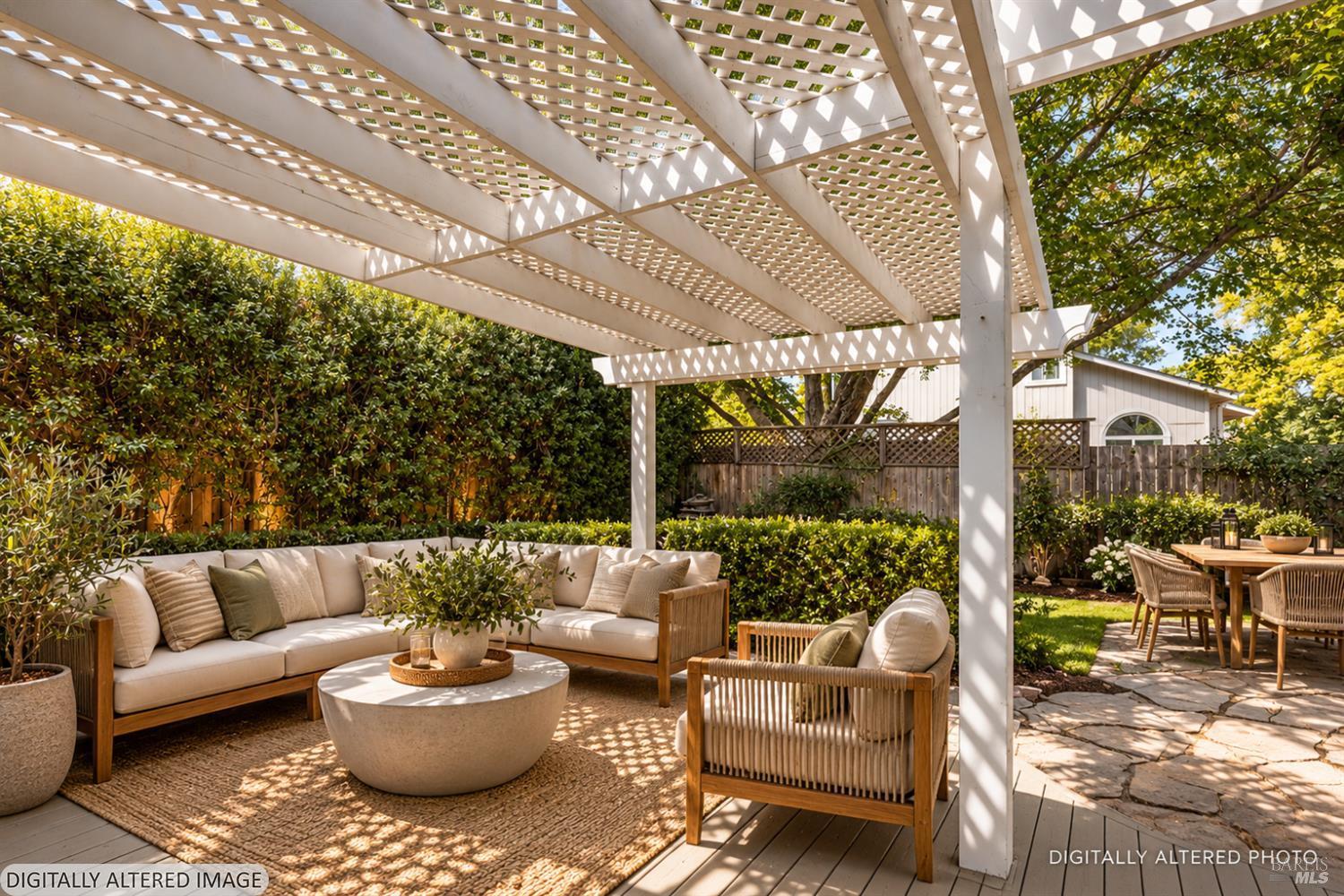 Backyard patio with a white pergola, cushioned lounge seating, dining table, and leafy privacy hedges.