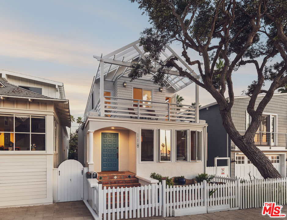 Two-story coastal home with a blue front door, upper balcony, white picket fence, and large front-yard tree.