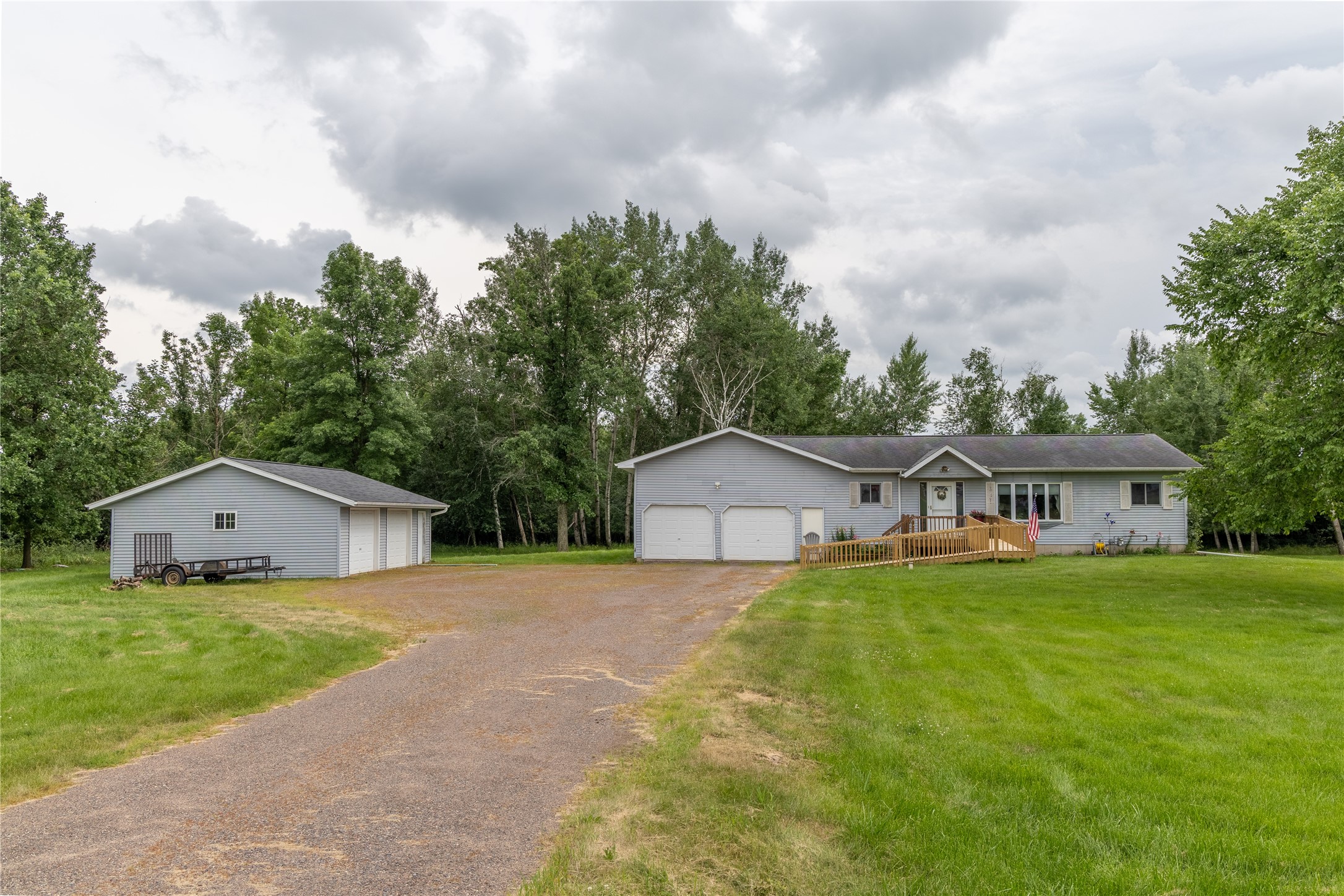 Single-story gray home with a wooden ramp, attached garage, separate outbuilding, and gravel driveway.