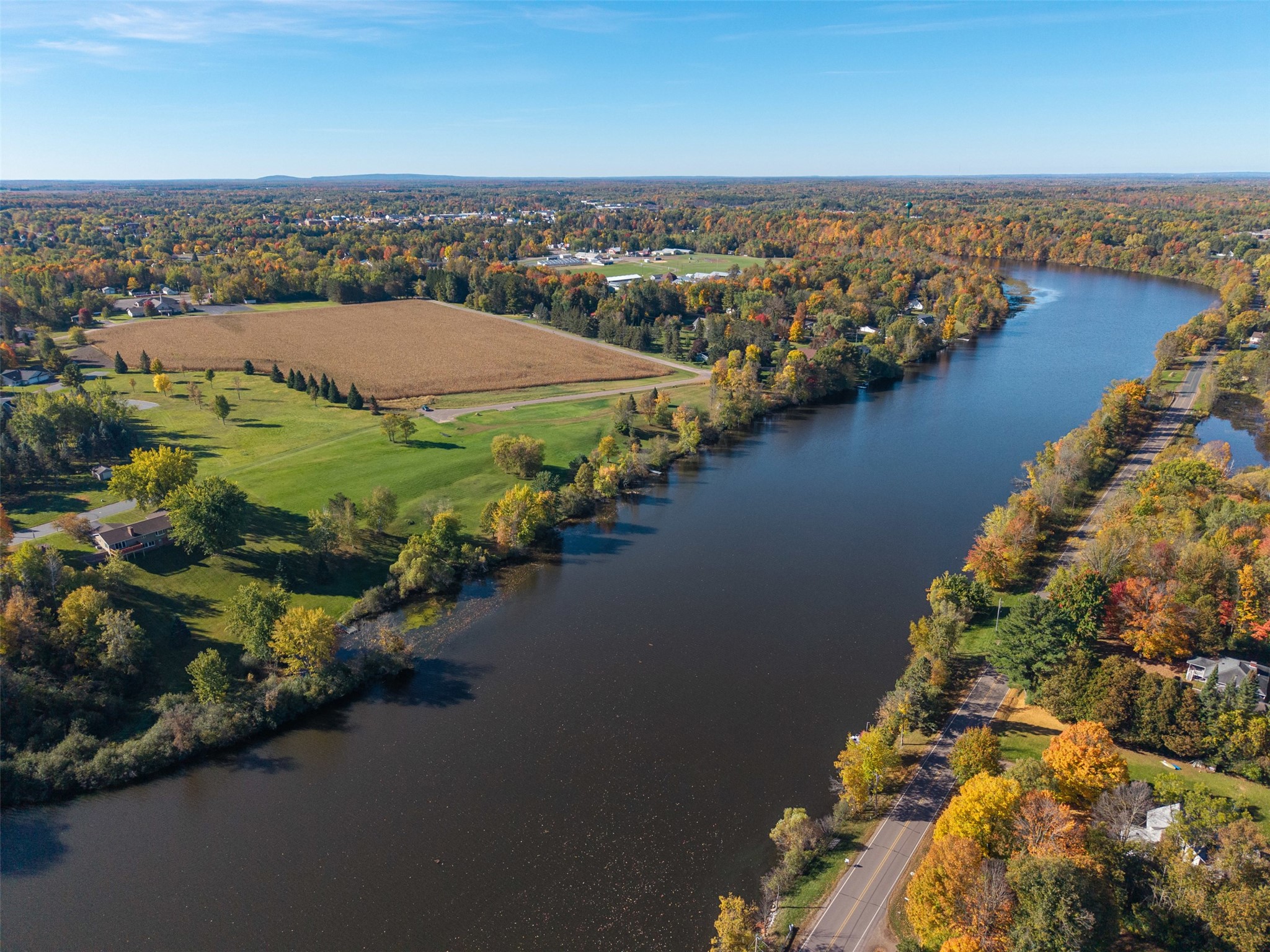Aerial view of a river bordered by autumn trees, homes, roads, and open fields.