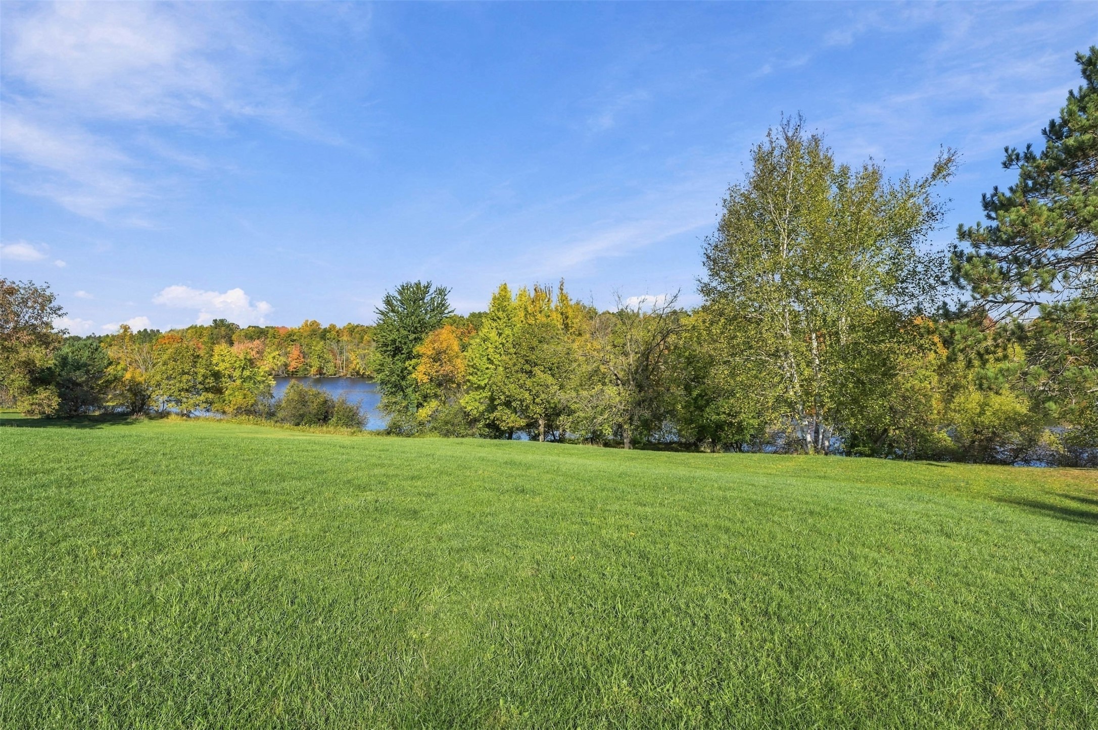 Grassy lot overlooking calm water and a tree-lined shoreline with autumn foliage.