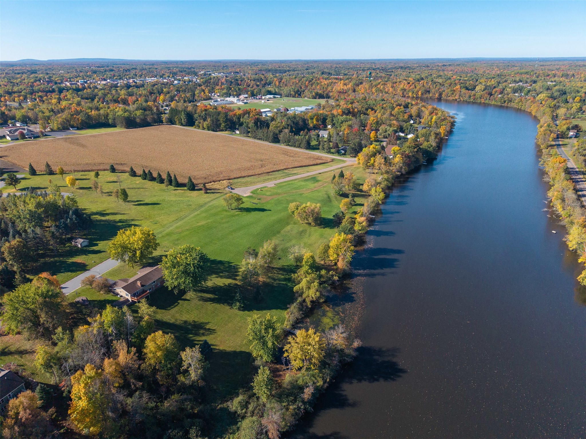 Aerial view of grassy riverfront land bordered by autumn trees, with a field and nearby homes.