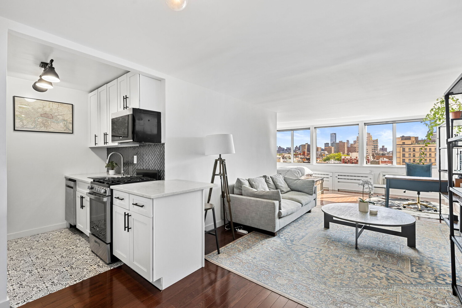 Open-plan apartment with a white kitchen, gray sofa, and city skyline views through large windows.