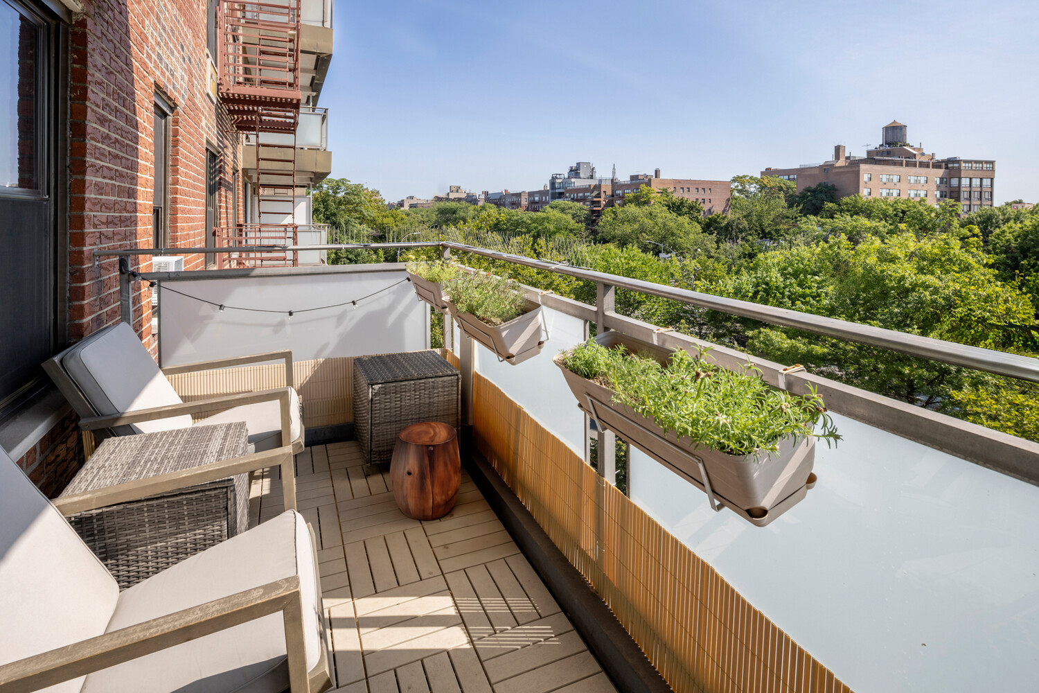 Furnished balcony with planter boxes overlooking leafy treetops and Brooklyn buildings.