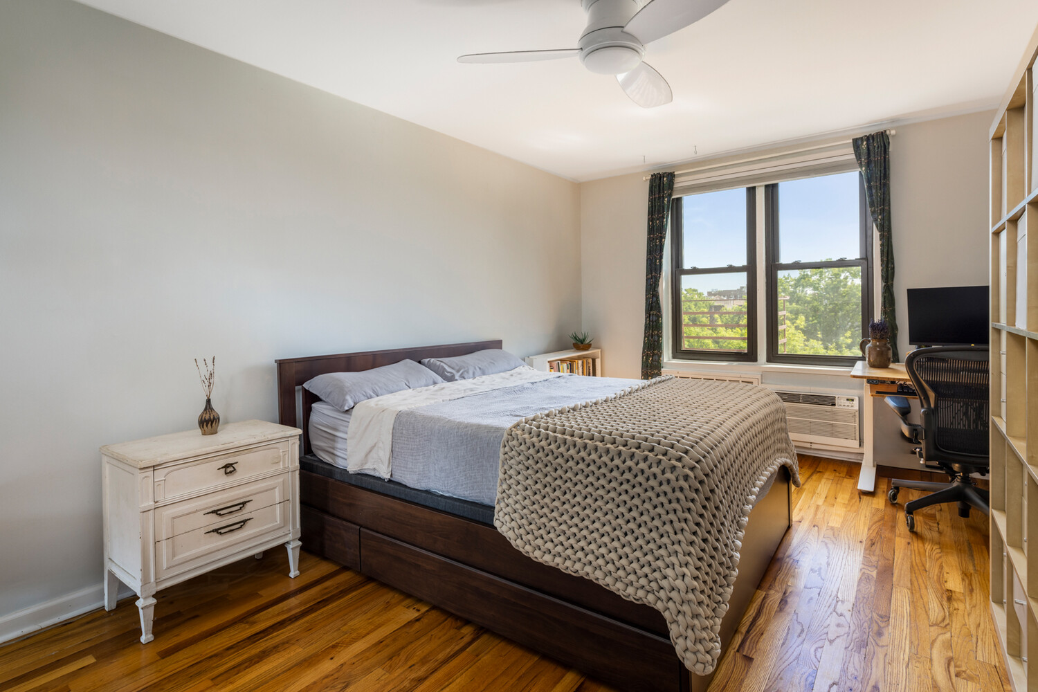 Bedroom with a dark wood bed, chunky knit blanket, hardwood floors, and large windows overlooking trees.