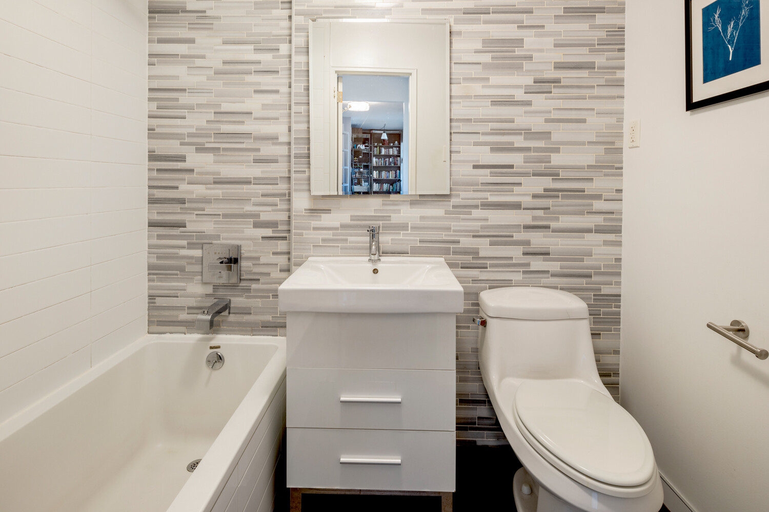 Bathroom with a bathtub, white vanity, and toilet against a gray mosaic tile wall.
