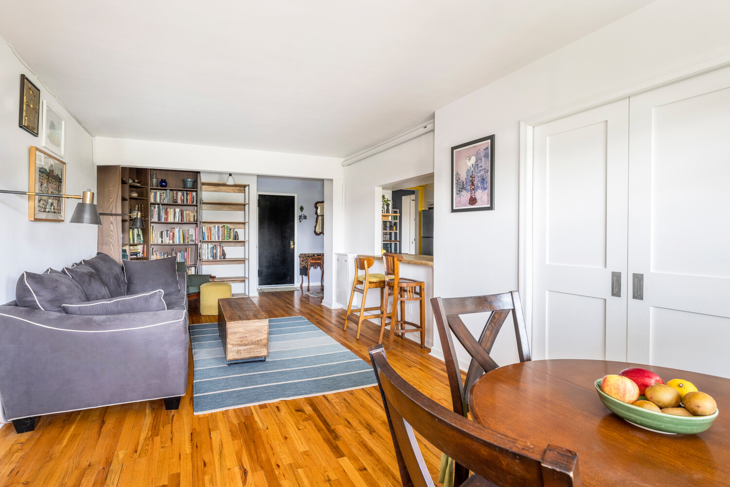 Furnished living and dining area with a gray sectional, bookshelves, hardwood floors, and a fruit bowl on the dining table.