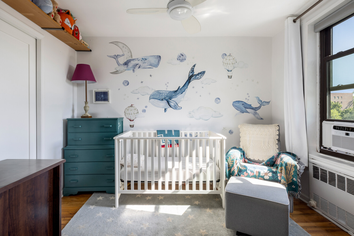 Nursery with a white crib beneath whale wall decals, a teal dresser, and a patterned armchair.