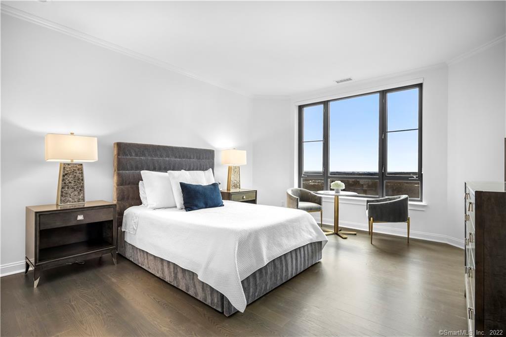 Staged bedroom with a gray upholstered bed, dark wood nightstands, and a window-side seating area.