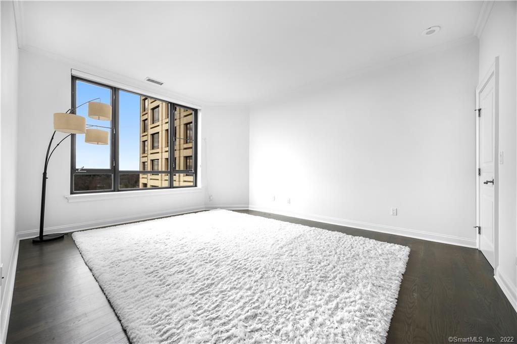 Mostly unfurnished bedroom with a large window, white rug, dark wood floor, and floor lamp.