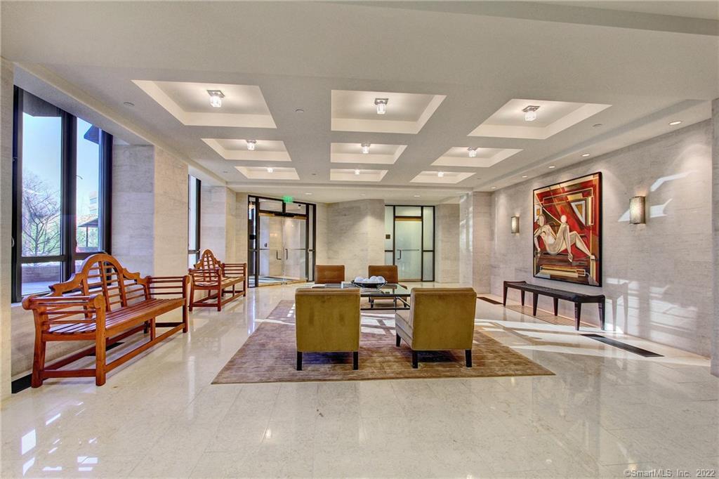 Condominium lobby with polished stone floors, tall windows, wood benches, and a central seating area.