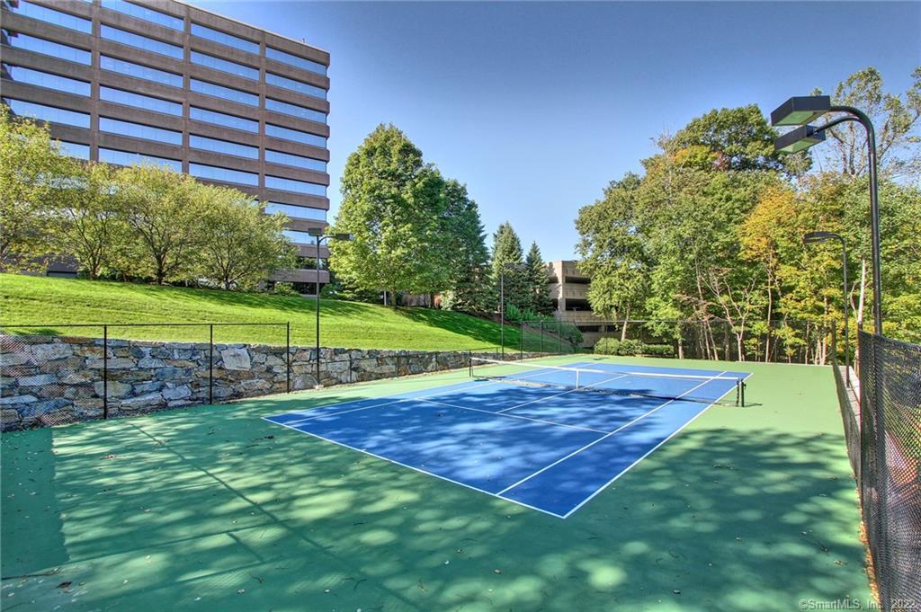 Outdoor tennis court beside a multi-story building, with a green-and-blue playing surface and surrounding trees.